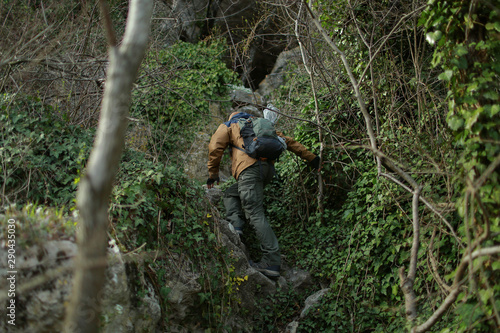 A man in a brown-and-blue windbreaker with backpack and rope climbs the slope of the Mangup plateau in Crimea towards the karst cave.