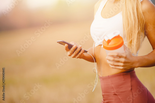 Girl uses phone for online training with connected headphones, in hand sports bottle for water