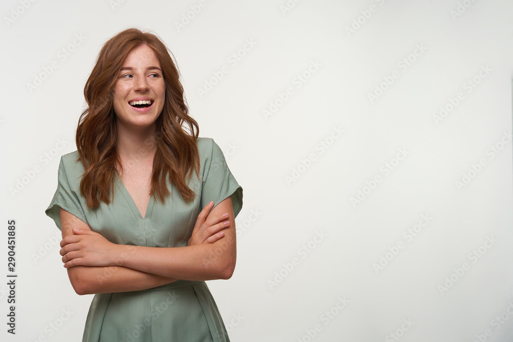Fototapeta premium Happy young pretty woman with red hair posing over white background with crossed arms, looking aside with wide and sincere smile, wearing vintage dress in pastel color