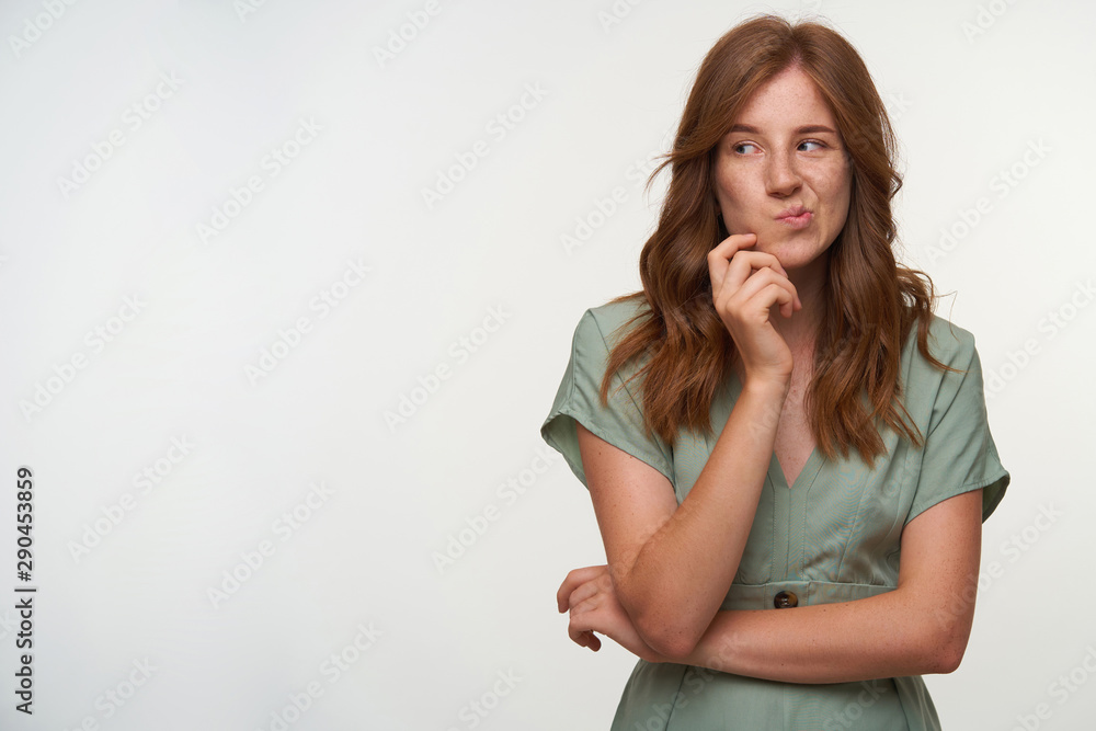 Studio close-up of pretty young redhead female holding her chin and biting cheek, looking puzzled aside, standing over white background Studio close-up of pretty young redhead female holding her chin and biting cheek, looking puzzled aside, standing over white background