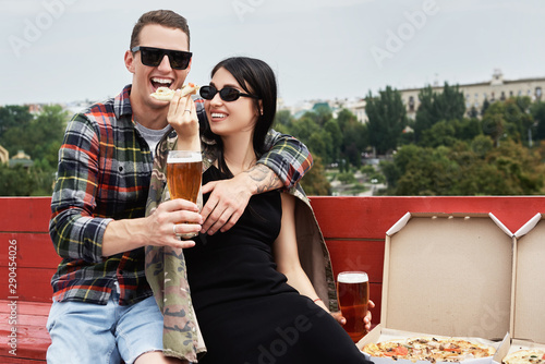 Happy smiling couple drinking beer and eating pizza at outdoor pub or bar on roof, copy space photo