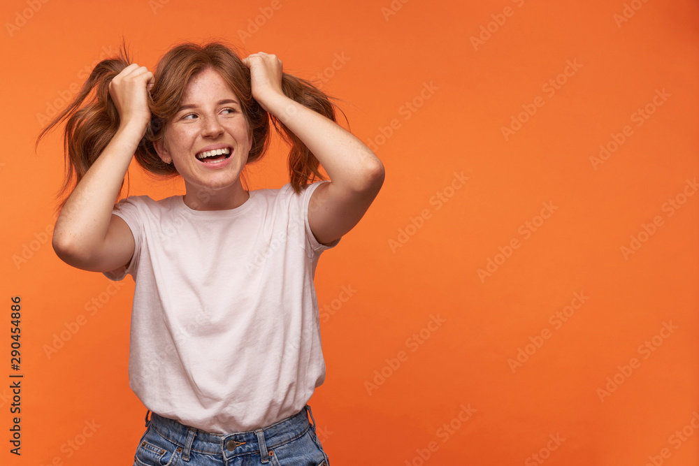 Happy young lovely woman with red hair standing over orange background in casual wear, looking aside with broad smile and holding tails Happy young lovely woman with red hair standing over orange background in casual wear, looking aside with broad smile and holding tails