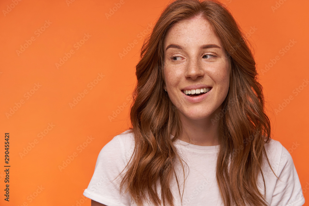 Indoor close-up with curly red haired cheerful female in white t-shirt, looking aside with charming smile, posing over orange background Indoor close-up with curly red haired cheerful female in white t-shirt, looking aside with charming smile, posing over orange background