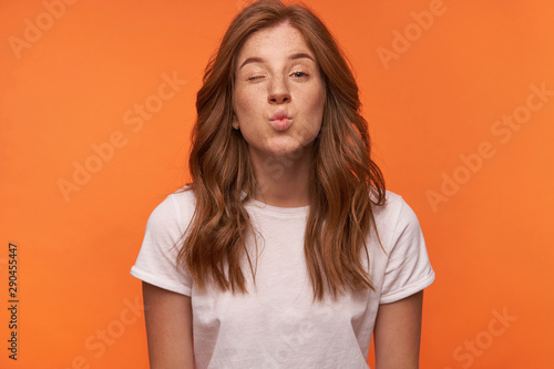 Positive young attractive female posing over orange background with hands down, winking and folding lips in air kiss, wearing casual wear