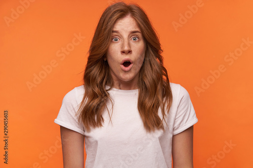 Studio shot of shocked pretty young woman with curly red hair standing over orange background, looking suprisedly to camera with wide mouth opened