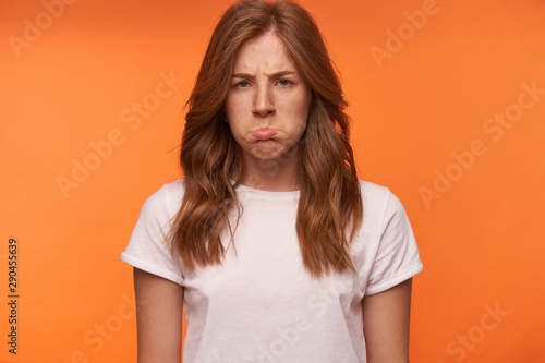 Studio close-up of charming offended young female posing over orange background, pouting and frowning with upset face, wearing casual wear