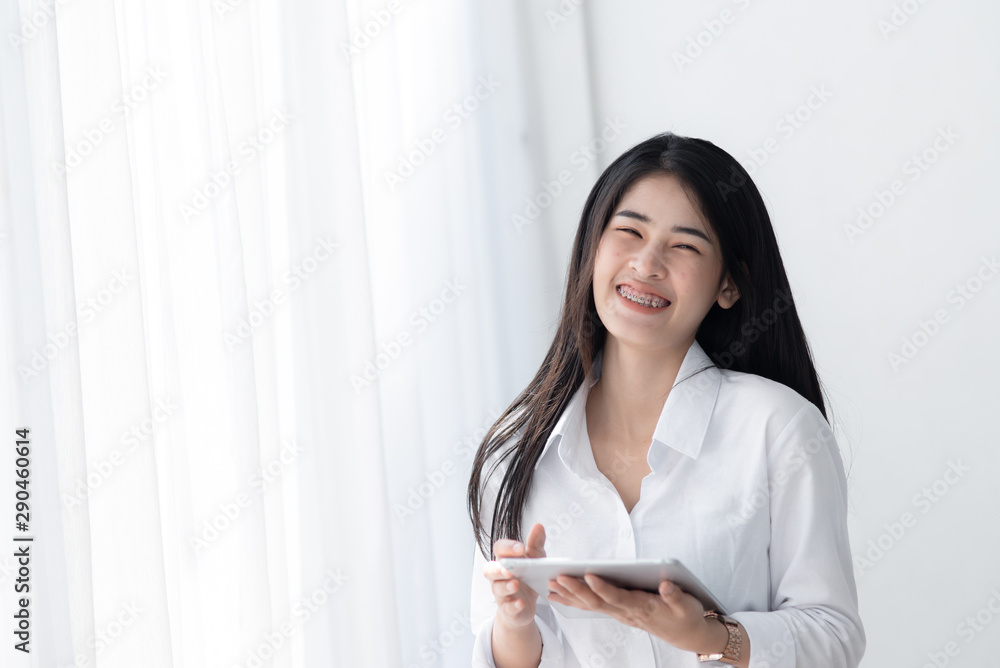 Beautiful Asian woman in white shirt using tablet near window in the morning. Beautiful Asian woman in white shirt using tablet near window in the morning.