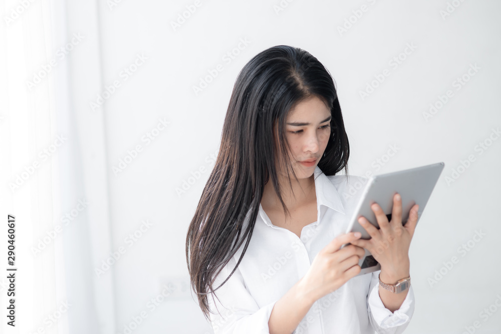Beautiful Asian woman in white shirt using tablet near window in the morning. Beautiful Asian woman in white shirt using tablet near window in the morning.