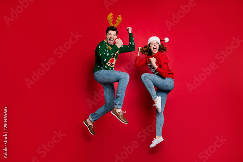 Full body photo of amazed jumping couple excited by x-mas prices wear ugly ornament jumpers and headwear isolated red color background