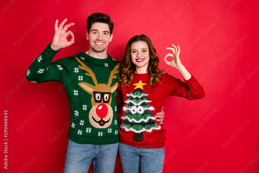Portrait of cheerful two people student with brunette wavy hair hug embrace show ok sign sales wear funny deer christmas tree pullover denim jeans isolated over red color background Portrait of cheerful two people student with brunette wavy hair hug embrace show ok sign sales wear funny deer christmas tree pullover denim jeans isolated over red color background