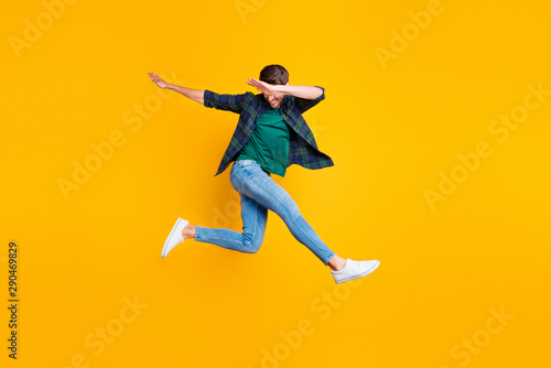Full body photo of cheerful content dream funky man jump have fun dance like true clubber enjoy summer vacation wear denim jeans sneakers checked green shirt isolated over yellow color background