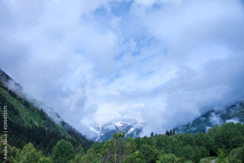 Beautiful panorama of the Caucasus mountains. Top of mountain range is covered in fog. Clouds lie on mountains. Summer day. Background image for travel and nature.