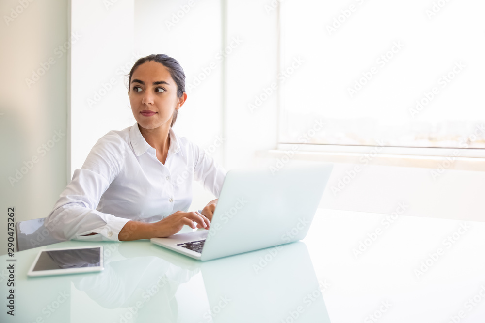 Confident female consultant working on computer. Young Latin businesswoman sitting at workplace, using laptop, looking away. Professional at workplace concept Confident female consultant working on computer. Young Latin businesswoman sitting at workplace, using laptop, looking away. Professional at workplace concept