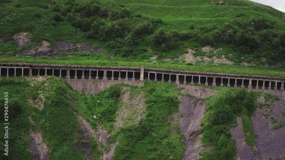 Crossover with trailer driving dangerous road on the side of the rocky cliff trough a tunnel carved with columns in the Caucasian mountain - Aerial drone view Crossover with trailer driving dangerous road on the side of the rocky cliff trough a tunnel carved with columns in the Caucasian mountain - Aerial drone view