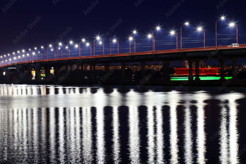 Fototapeta premium The night bridge with beautiful lanterns is reflected in the water on the Dnieper River, in the Dnepropetrovsk city, Ukraine. Evening river landscape in a big city.