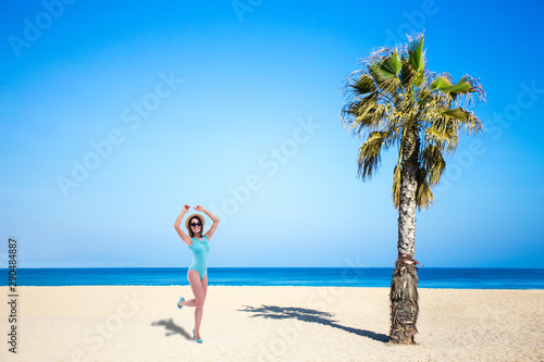 summer and travel concept - young beautiful woman in swimsuit dancing at the beach near the palm tree - copy space over blue sky