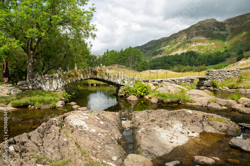 Slaters Bridge in the Lake District National Park with fells/mountains in the background and river and rocks in the foreground