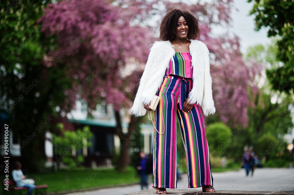 Fashionable african american woman in pink striped jumpsuit with fluffy faux fur coat posed at spring bloom street.