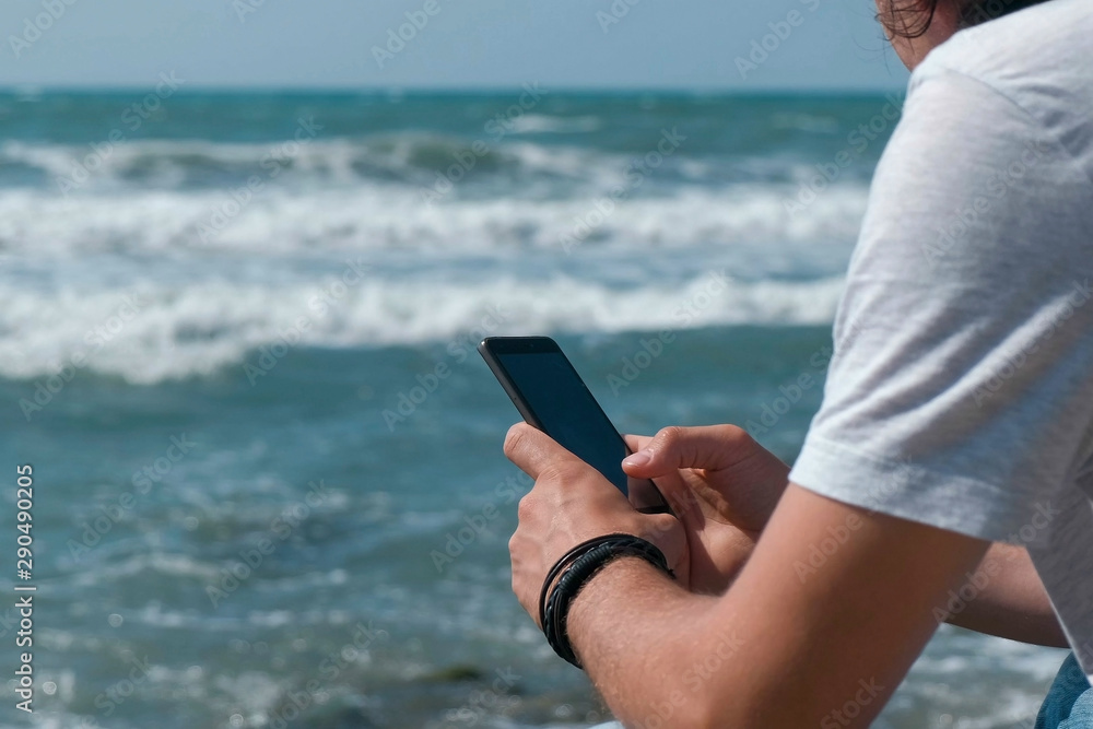 Young man types a message on mobile phone sitting on the big stone on sea beach. Hands close-up