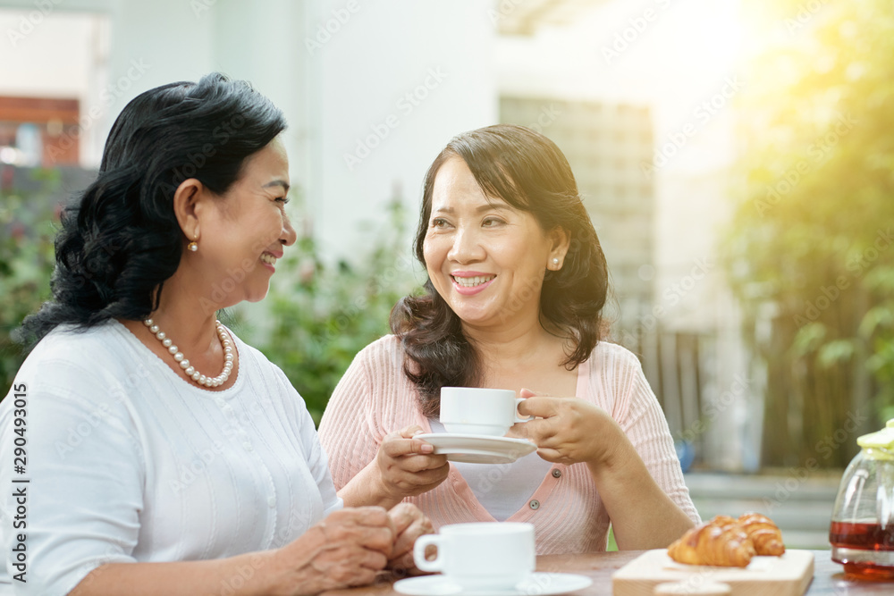 Two Asian smiling women sitting at the table drinking coffee and talking to each other during their breakfast in outdoor cafe Two Asian smiling women sitting at the table drinking coffee and talking to each other during their breakfast in outdoor cafe