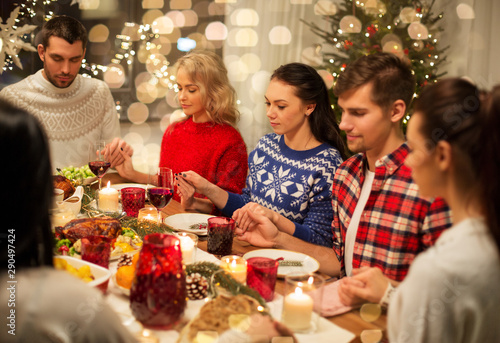 holidays and thankfulness concept - friends holding hands and praying while having christmas dinner at home