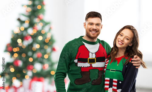 winter holidays, celebration and people concept - portrait of happy couple in ugly sweaters at home over christmas tree lights background