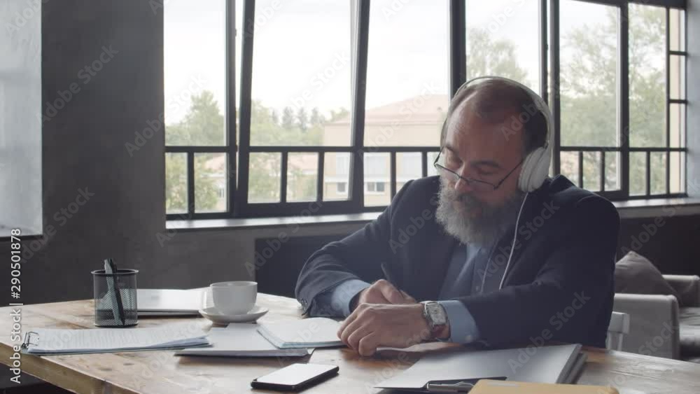 Tracking right of senior male writer sitting at desk, writing in notebook while listening to music through headphones Tracking right of senior male writer sitting at desk, writing in notebook while listening to music through headphones