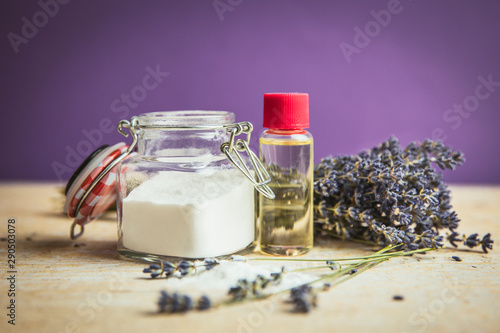 Olive oil and baking soda exfoliating scrub concept. Homemade natural beauty cosmetics. Jar with baking soda powder and bottle with olive oil, lavender bouquet on purple background.