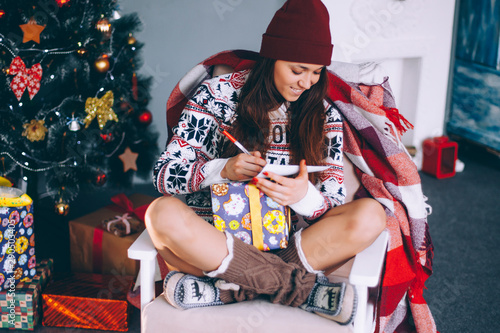 Portrait of a beautiful young woman near a New Year tree with gifts. A young woman dressed in a New Year's sweater writes a letter to Santa Claus.