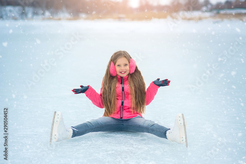 Cheerful Winter Ice skating Girl having fun on ice skate rink outdoors. Cute photo of young smiling girl sitting on the ice with open hand. Ice skating rink