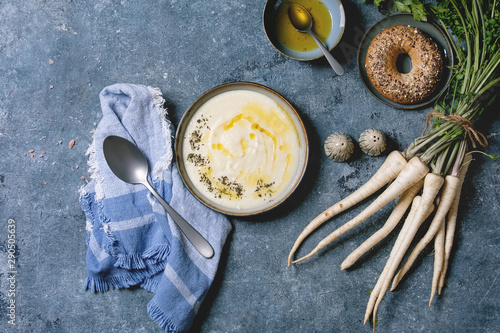 Parsnipcream soup in ceramic bowl with butter sauce, bagel bread, bundle of fresh parsnip and herbs over blue texture background. Flat lay, space © Natasha Breen