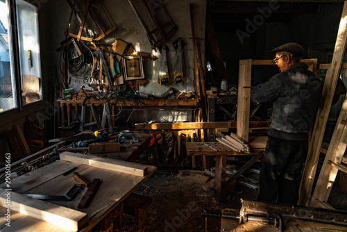 Old master carpenter in grey clothes is getting ready for work in his home workshop, many tools hang on the wall