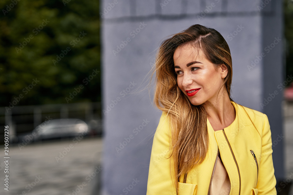 Fototapeta premium Portrait of a young beautiful laughing woman with long hair and a yellow jacket against the background on a cloudy day. Close-up.