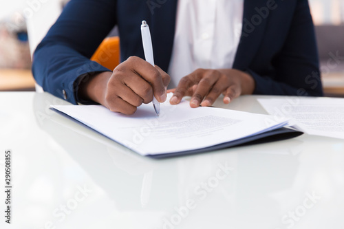 Businesswoman signing contract. African American business woman sitting at table in office, holding pen and writing in document. Legal expertise concept photo