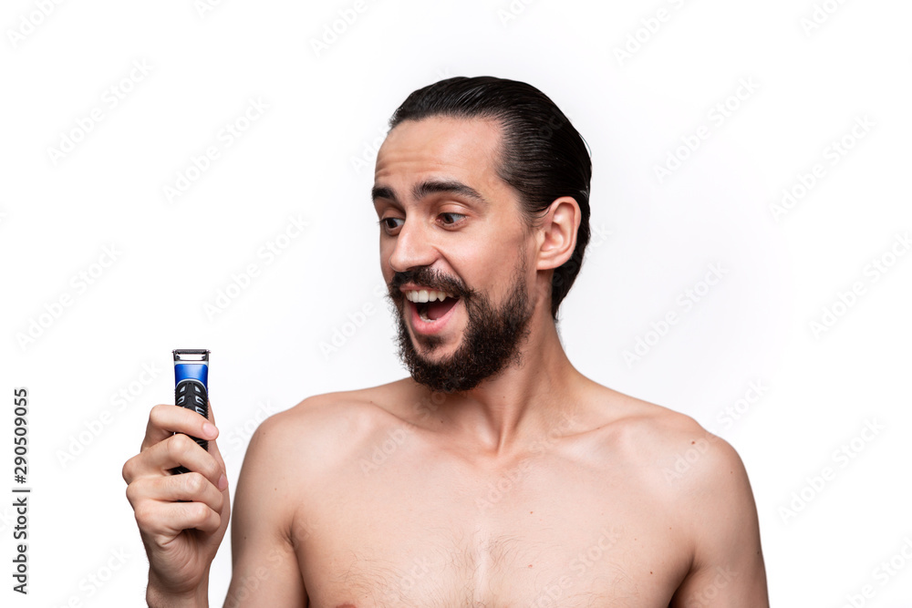 Fototapeta premium Smiling bearded man with mustache looks at the electric shaver standning bare isolated over white background. Concept of morning treatment and shaving.