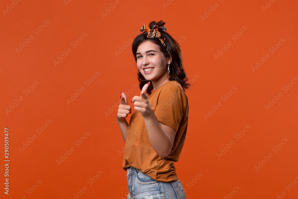 Pretty brunette woman in a t-shirt and beautiful headband pointing with her fingers at the camera isolated over orange background. Place for ad. Pretty brunette woman in a t-shirt and beautiful headband pointing with her fingers at the camera isolated over orange background. Place for ad.