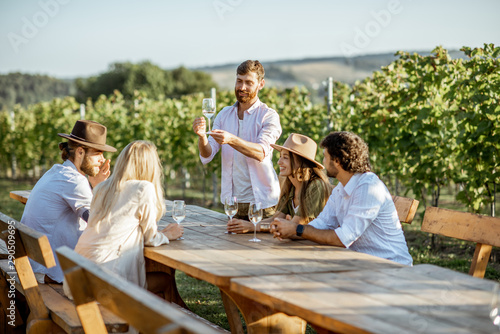 Group of a young people drinking wine and talking together while sitting at the dining table outdoors on the vineyard on a sunny evening photo