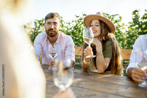 Group of a young people drinking wine and talking together while sitting at the dining table outdoors on the vineyard on a sunny evening