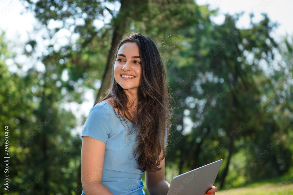 Obraz premium Young brunette student in a blue dress sitting with a laptop in a park. Distance learning