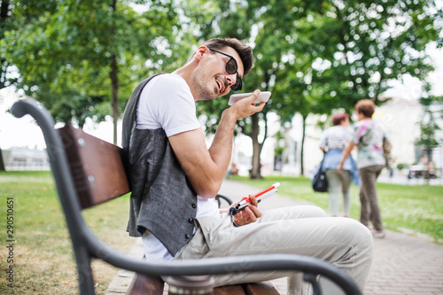 Young blind man with smartphone sitting on bench in park in city. © Halfpoint