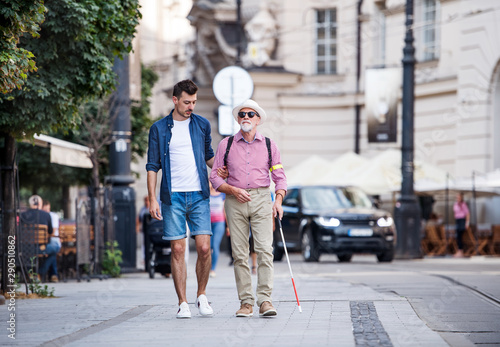 Young man and blind senior with white cane walking on pavement in city.