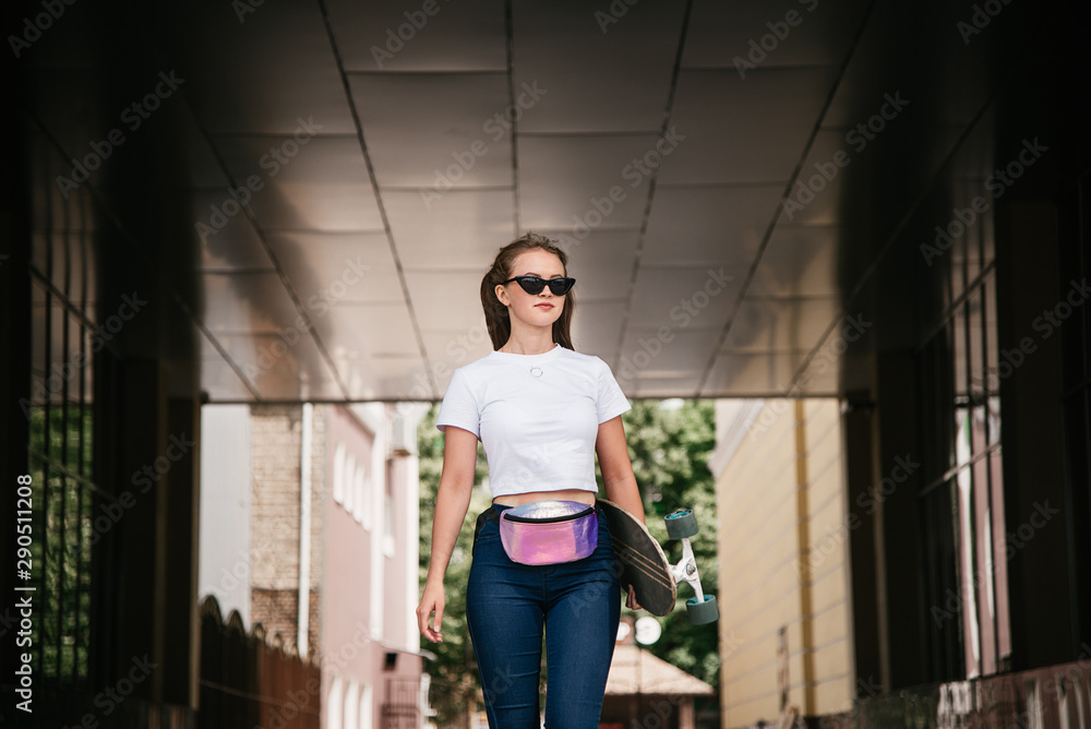 A young girl in sunglasses, white t-shirt and blue jeans is walking down the street with skate board in her hands. A young girl in sunglasses, white t-shirt and blue jeans is walking down the street with skate board in her hands.
