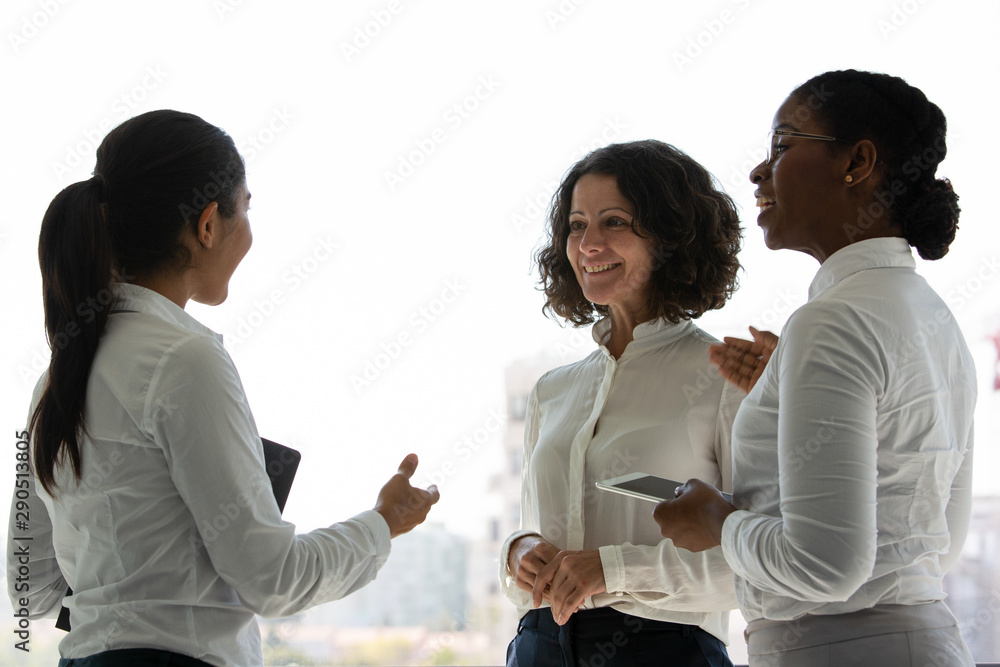 Team of happy female colleagues chatting near office window. Businesswomen standing together, talking, gesturing and smiling. Business networking concept Team of happy female colleagues chatting near office window. Businesswomen standing together, talking, gesturing and smiling. Business networking concept