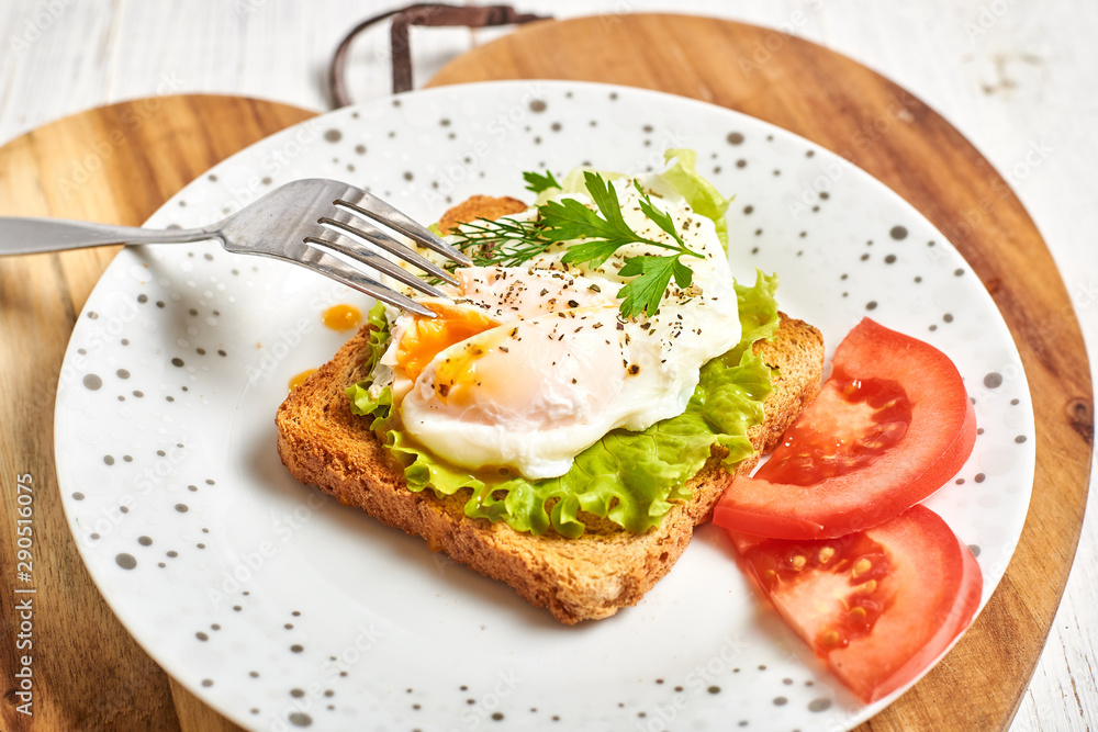 Poached egg, benedict on toasted white bread toast with salad and spices on a plate on a light wooden background.