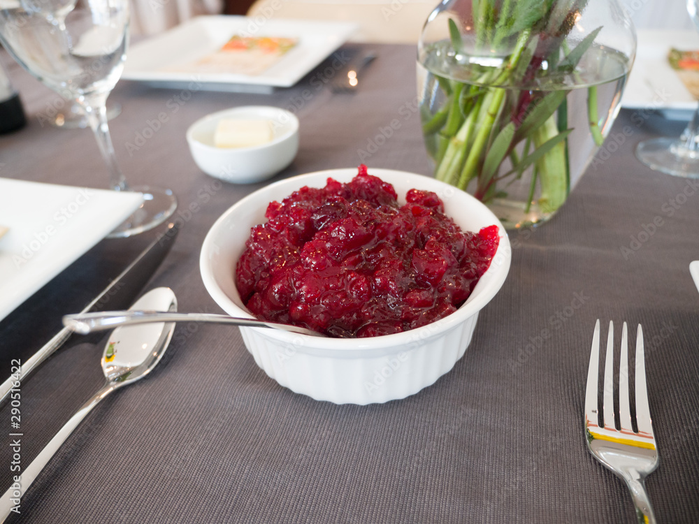 A small white corrugated ceramic serving bowl full of bright red chunky cranberry sauce set between two table settings with silverware, plates and flower vase in background for Thanksgiving dinner. A small white corrugated ceramic serving bowl full of bright red chunky cranberry sauce set between two table settings with silverware, plates and flower vase in background for Thanksgiving dinner.