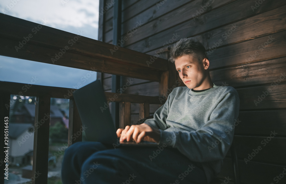 Handsome guy uses a laptop in the evening on the balcony.Portrait of a freelancer.Portrait of young man working at laptop at night on balcony in apartment, looking at laptop screen with serious face