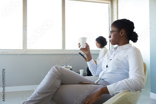 Office employees drinking coffee in office lounge. Young business woman sitting in armchair, holding and looking at disposal cup. Work balance concept