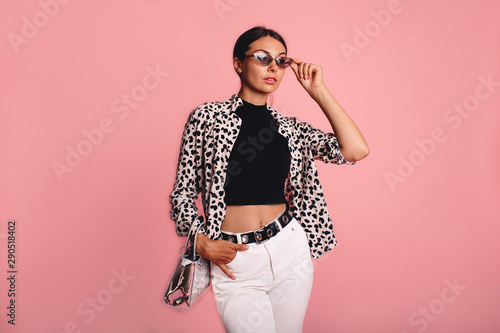 Fashion photo of a beautiful young woman in a casual summer look with animal print shirt, bag and white jeans posing over pink background. Fashion photo, copy space