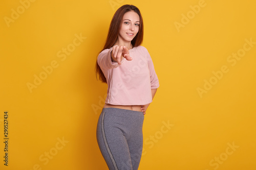 Portrait of young Caucasian athletic girl pointing at camera, looking smiling at camera, wearing stylish sporty clothing, model posing isolated over yellowstudio background.Fitness and sport concept.