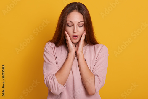 Close up portrait of surprised beautiful girl holding her hands near cheeks and keeps mouth opened, attractive lady being in amazement, posing isolated over yellow background. People emotions concept. photo
